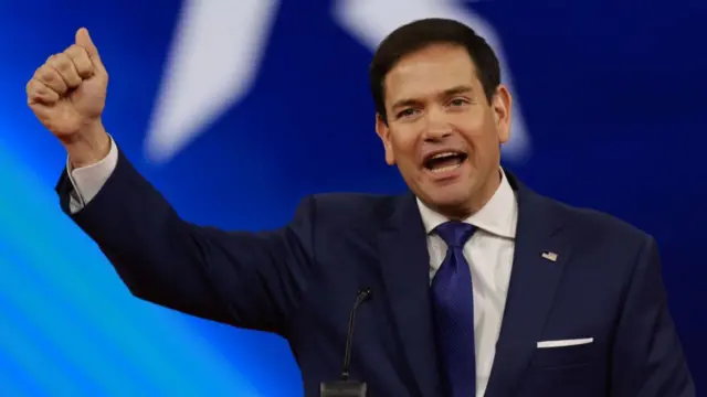 Marco Rbuio, wearing a blue suit with a blue tie, lifts his fist and smiles