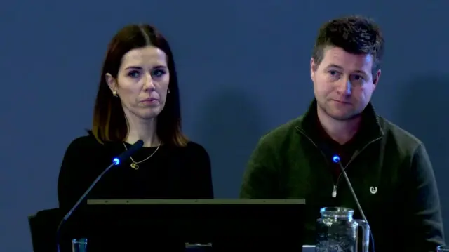 Ronan McGrory sits beside a woman. He is on the right, sitting behind a desk with two microphones in front of him. he is wearing a green top and has short brown hair