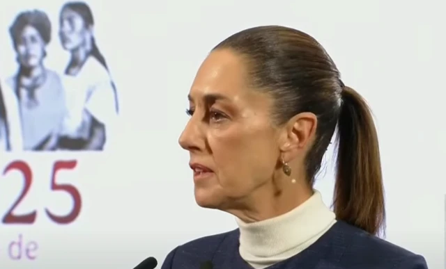 Sheinbaum stands in front a white screen. She wears a ponytail in her hair.