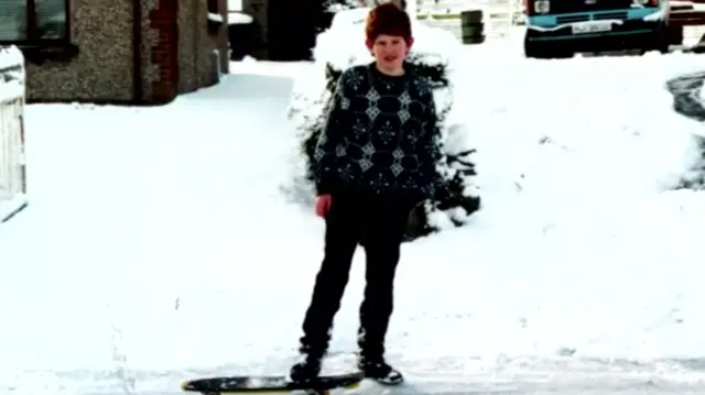 Aiden Gallagher stands in a snow street, he is a child and his foot is on a skateboard. He is wearing black.
