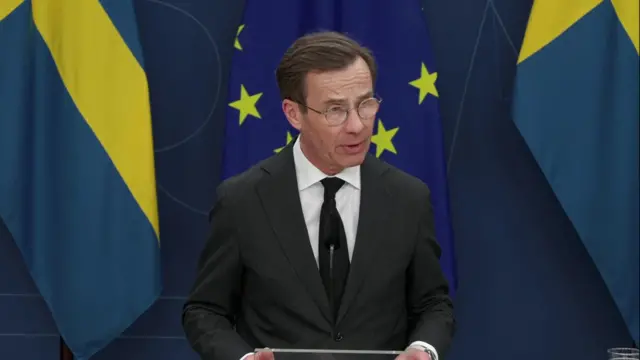 Swedish prime minister talking into a microphine with the Swedish and European Union flags behind him