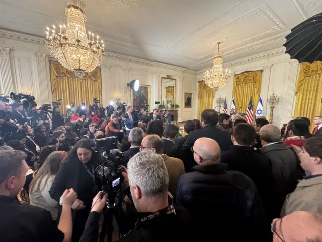 The East room of the White House, with dozens of reporters gathered there with cameras and lights. Israeli and US flags are visible where Trump and Netanyahu will be speaking later.