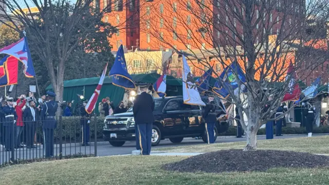 A black armoured SUV arriving at the white house. Members of the US military in uniform are lining the road it is driving on, waving a variety of flags from around the US.