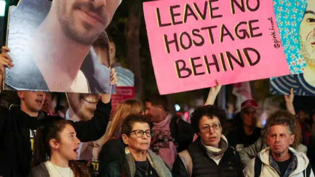 A group of people hold posters and images. One reads 'Leave no hostage behind'