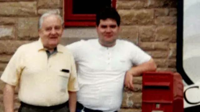 Fred and Bryan White stand side by side - Fred in a yellow shirt and Bryan in a white T-Shirt. Bryan is leaning on a red post box.
