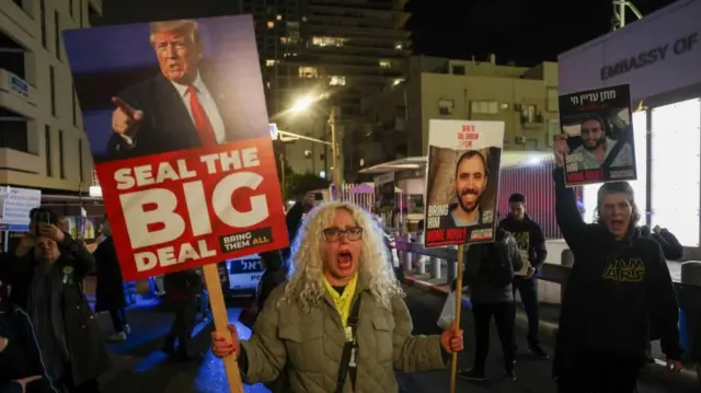 A woman holds a placard featuring an image of Trump with the slogan 'Seal the Big Deal, Bring them all'