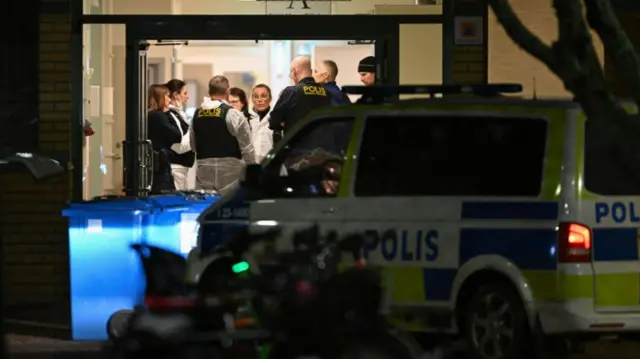 Police at the school where the shooting took place. Some wear protective suits, and a police car is parked outside the entrance