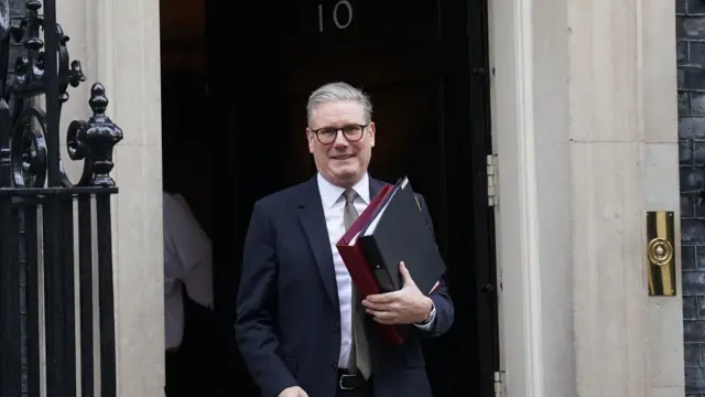 Keir Starmer smiles as he leaves No 10, wearing a dark suit and clutching red and black folders