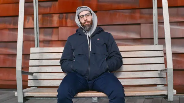 Tariq Luthun sitting on a bench, he has a large beard, has glasses on and wearing a hoodie