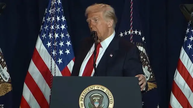 Trump standing facing slightly sideways at the lectern speaking into a microphone in front of two American flags