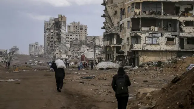 Two people walking towards many ruined buildings that lay in front of them