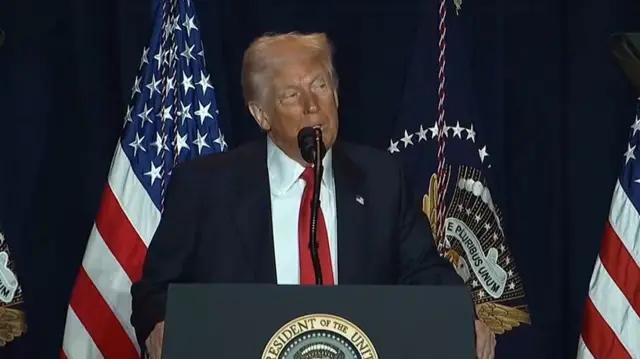 Trump speaking in front of an American flag into a microphone at a lectern