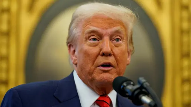 U.S. President Donald Trump in blue suit and red tie speaking into microphone with gold background