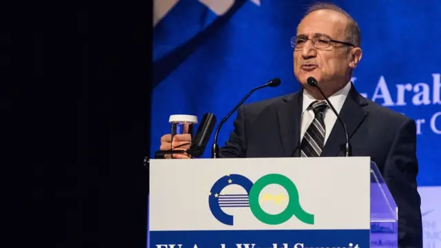 al-Anani speaking at a lectern into a microphone. A glass of water is placed in front of him