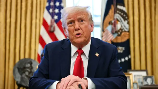 Donald Trump sitting down at his wooden desk in the Oval Office in dark suit and red tie, Family photos are visible to his left on a small table