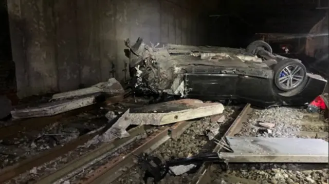 A burnt out car lies upside down across railways lines, with debris all around