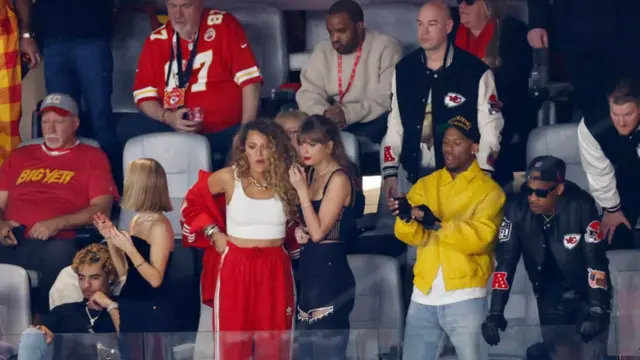 Taylor Swift, wearing a black outfit, and Blake Lively, wearing a red and white track pants and white tank top, watch in the stands of the Super Bowl in 2024