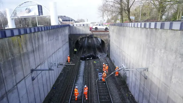 A wrecked car lies upside down across two train tracks. The bridge overhead is missing a panel where the car appears to have smashed through and plunged down. Emergency workers assess the scene