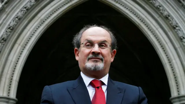 Salman Rushdie is seen in front of a courthouse. He has a red tie and blue suit on. He wears glasses and has grey hair.