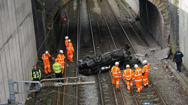 Emergency workers assess the scene as the wrecked car lies upside down across two tracks