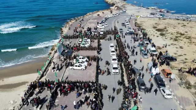 Long queues of people stand near lines of Red Cross vehicles along a coastline