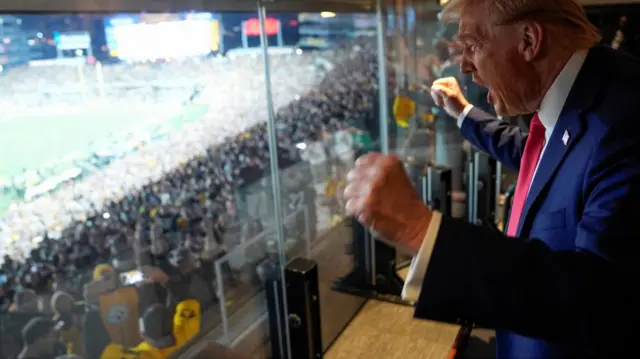 Donald Trump cheers with his fists clenched while watching a football game. He wears a blue suit and red tie, and his mouth is open as he shouts a cheer.