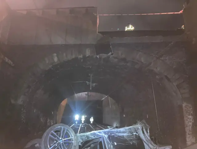 A burnt out car lies upside down on a railway track in this image captured from the railway line. People in hi vis outfits can be seen approaching it, under the railway bridge.