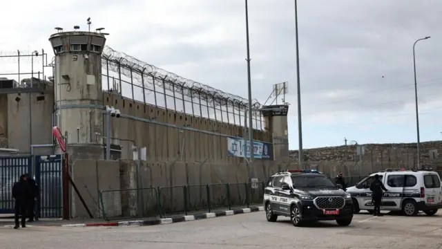 The Ofer prison in the occupied West Bank is shown. It is built of grey concrete, with a tall fence and barbed wire wrapped around it. Two police vehicles are parked out front, one is black and one is white. Both are SUV-style vehicles. Two officials, both dressed in dark clothes, mill in the background. The prison walls cover most of the photograph.