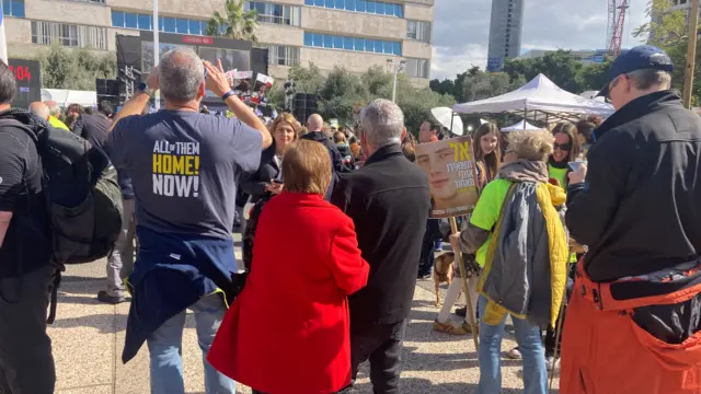 A group of people dressed in brightly coloured red and green clothing stand in hostages square. one man wears a navy shirt that says 'all of them home now'. They are watching large screens showing a newscast of the hostages being released. Some hold placards with pictures of their loved ones.