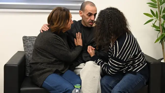 Eli Sharabi sits with his mother and sister. One of the women has short brown hair and the other has dark, long, curly hair. Eli Sharabi has a shaved head.