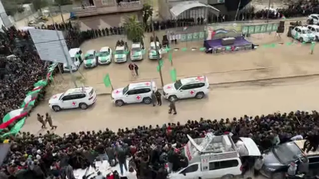 Three white Red Cross vehicles arrive at hostage release staging area. There is a huge crowd surrounding the cars and a purple stage in the background