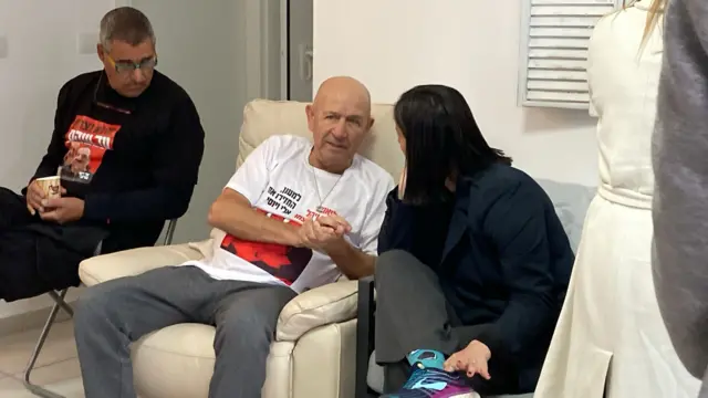 Jacob Srdnayof sits in a white leather armchair with two relatives. He is bald and in his mid-to-late 50s. He is wearing a white t-shirt with a red photo on it that is partially obscured by his arm. His relatives are speaking to him, there is a man and a woman, both with dark hair. Both relatives are listening as Jacob speaks.