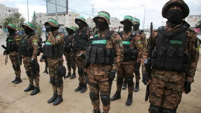 Hamas fighters stand in rows in formation in Deir al-Balah. There are five fighters in each row, about three rows deep. They're dressed in military fatigues with masks over their faces. They are wearing bucket hats branded with Hamas' flag. They're standing in what appears to be a town square.