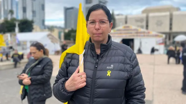 Keren Redlich is wearing a black puffer jacket and carrying a yellow flag. She has a yellow ribbon pinned to her jacket. She has dark hair and glasses. She is standing in hostages square.