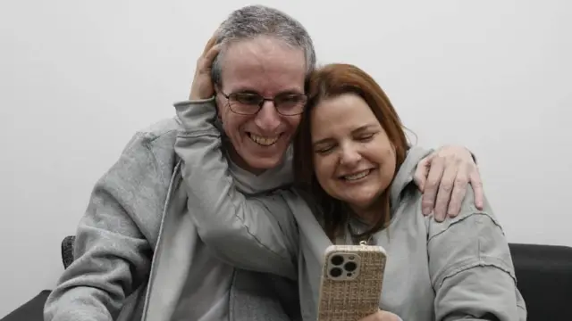 An older man, who looks quite thin, smiles as he wraps his arm around a woman sitting next to him on a couch. She is smiling broadly as the pair look at a phone, in what appears to be a video call