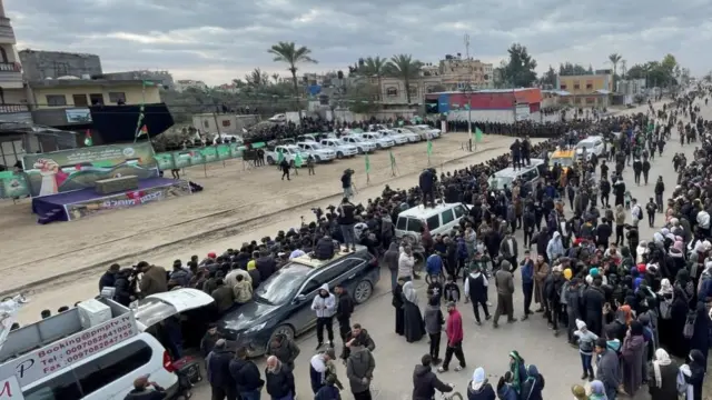 Groups of people gather waiting for Israeli hostage release. There is a stage and several cars.