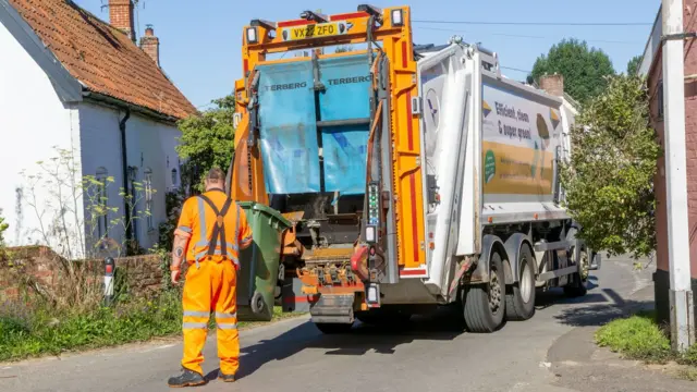 In a Suffolk country lane a council worker dressed in reflective orange guides a green wheelie bin into a waste collection lorry.