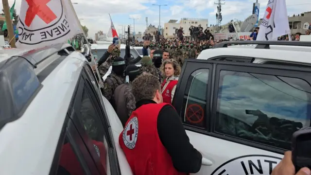 ICRC members stand between ICRC cars for transporting the Israeli hostages into. Hamas members stand in a crowd near the cars.