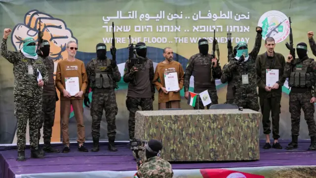 Or Levy, Ohad Ben Ami, and Eliyahu Sharabi stand on a stage alonside Hamas gunmen