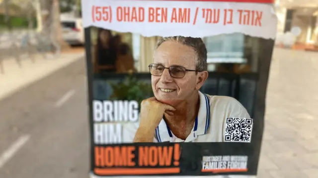 A poster in hostages square shows Ohad Ben Ami. The poster reads "bring him home now!" and shows a portrait of Ben Ami. He is wearing a white polo shirt and square-framed glasses and is resting his head on his hand. He's smiling. The poster has orange and white writing, and a QR code.