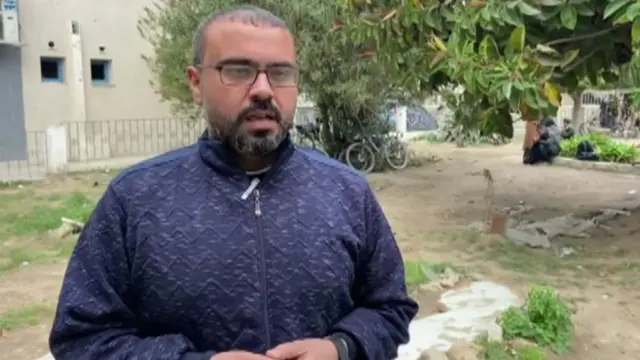 Hamas Qassem. He is wearing square, black-rimmed glasses and has short black hair and a black beard. He is wearing a black zip-up jumper.