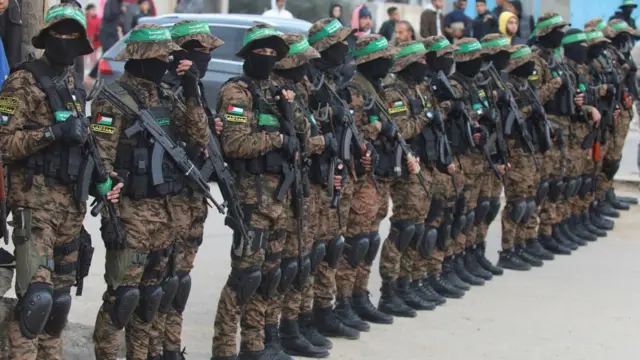 Hamas fighters stand in a row. They are dressed in army fatigues and wearing knee pads and hats that come down low over their eyes. They are wearing bullet proof vests and carrying guns. Green flags are wrapped like bandanas around their hats. There are about 15 to 20 fighters in the image.