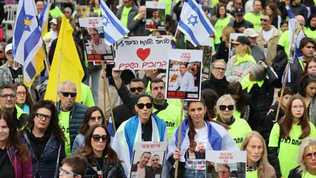 A large crowd of people dressed in bright green and wrapped in Israeli flags stand in Hostages square. they're carrying yellow flags and israeli flags, and holding placards of their loved ones. There are about 100 people in the image.