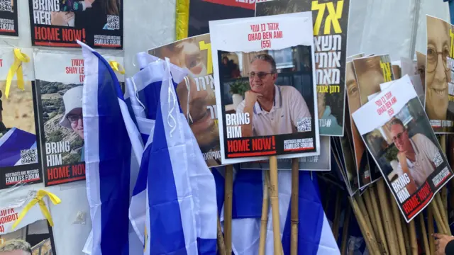 Several protest signs are leaning against a wall with Israeli flags on it. They feature photos of Ohad Ben Ami, who is wearing a whie polo shirt and glasses. He is resting is head on his hand. Yellow ribbons that are used by the hostage support organisations are tied to the placards.