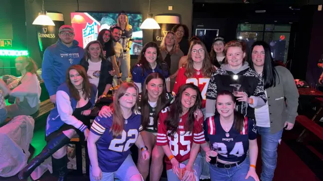 A group of women in American football tops of different teams at an Irish Bar with a TV in the background and Guinness ads on the wall.