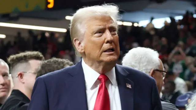 Donald Trump wearing a suit and red tie with people in the background