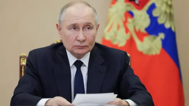 Putin sits at a desk and holds a piece of paper. There is a Russian flag behind him