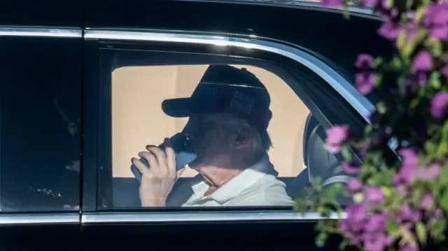 Donald Trump inside a car wearing a red Maga cap and holding a phone to his mouth