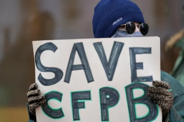 A man with sunglasses and a face covering holds a sign that says "Save CFPB"