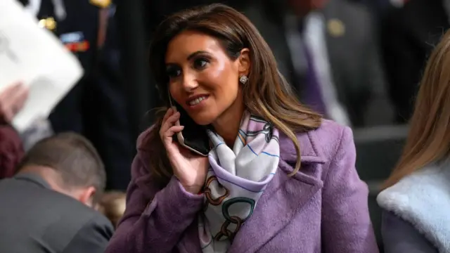 Alina Habba talking on the phone. She has long brown hair, brown eyes, and is wearing a lilac-coloured coat.